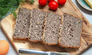 Chleb gryczany bezglutenowy przepis: kromki ciemnego chleba z ziarnami na desce, obok pomidorki, jajko i nóż.