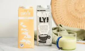 Dwa kartony mleka owsianego, w tym Oatly Barista Edition, idealne do kawy. Obok szklanka z zielonym napojem.