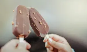 Dwie ręce trzymają czekoladowe lody na patyku z orzechami. Idealne na lato, wegańskie lody gdzie kupić?