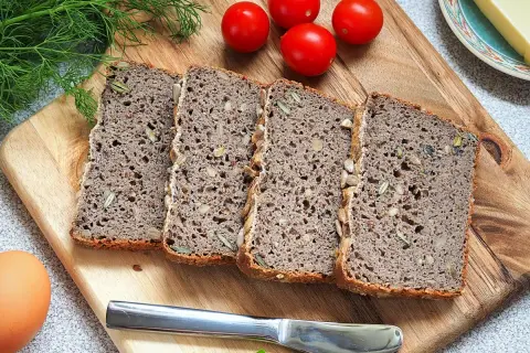 Chleb gryczany bezglutenowy przepis: kromki ciemnego chleba z ziarnami na desce, obok pomidorki, jajko i nóż.