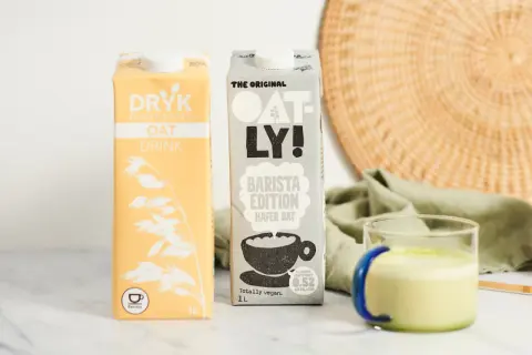 Dwa kartony mleka owsianego, w tym Oatly Barista Edition, idealne do kawy. Obok szklanka z zielonym napojem.