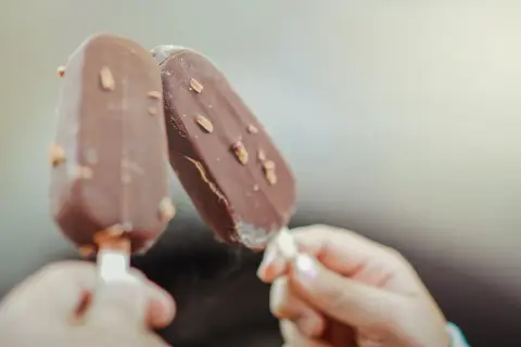 Dwie ręce trzymają czekoladowe lody na patyku z orzechami. Idealne na lato, wegańskie lody gdzie kupić?