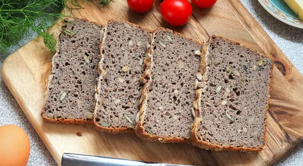 Chleb gryczany bezglutenowy przepis: kromki ciemnego chleba z ziarnami na desce, obok pomidorki, jajko i nóż.