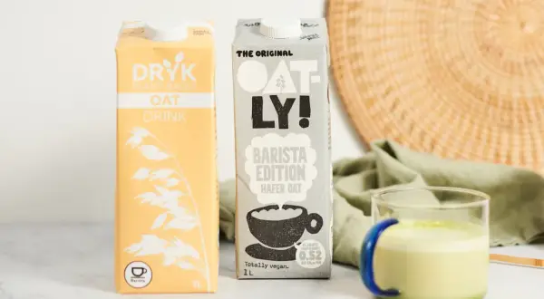 Dwa kartony mleka owsianego, w tym Oatly Barista Edition, idealne do kawy. Obok szklanka z zielonym napojem.