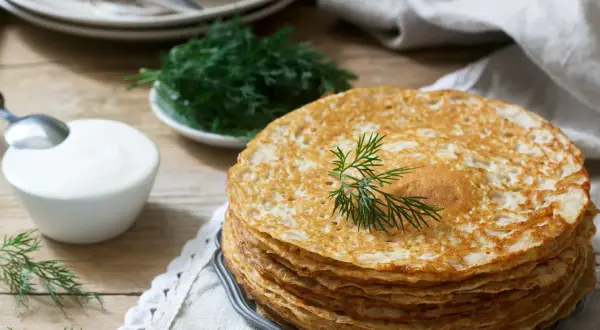 Stos naleśników bezglutenowych, ozdobiony gałązką koperku, obok miseczki śmietany i świeżego koperku. Idealne na śniadanie.