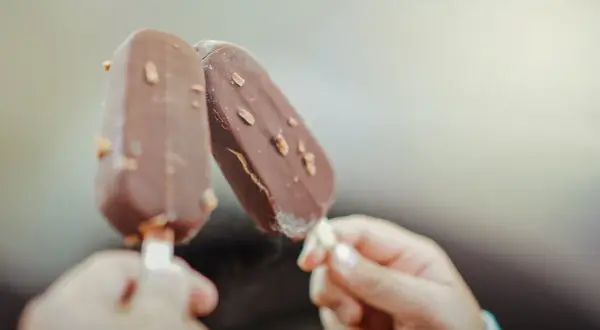 Dwie ręce trzymają czekoladowe lody na patyku z orzechami. Idealne na lato, wegańskie lody gdzie kupić?