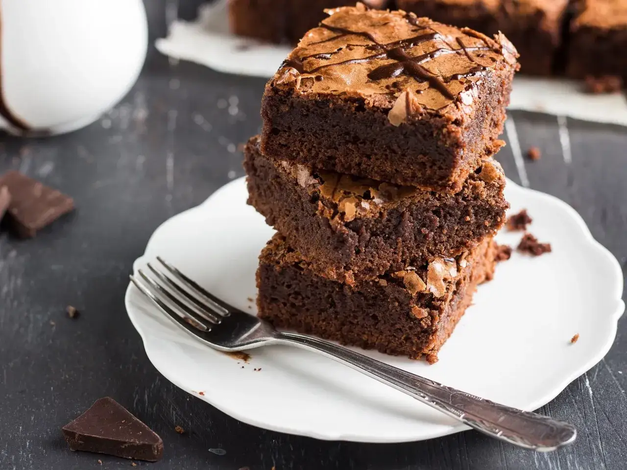 Pyszne, wilgotne brownie ułożone na talerzyku, obok widelca i kawałków czekolady. Idealny przepis na zdrowe ciasto.
