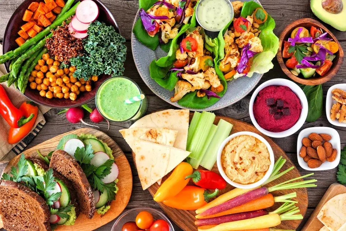 Kolorowe dania na drewnianym stole pokazują, co jeść na diecie wegańskiej: sałatki, wrapy z kalafiora, hummus, warzywa i smoothie.